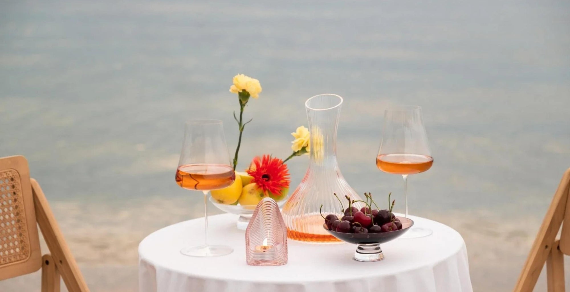 Two NUDE Stem Zero Volcano white wine glasses of rosé with a NUDE Vini wine carafe, Mist votive and Silhouette bowl on a white table by the water.