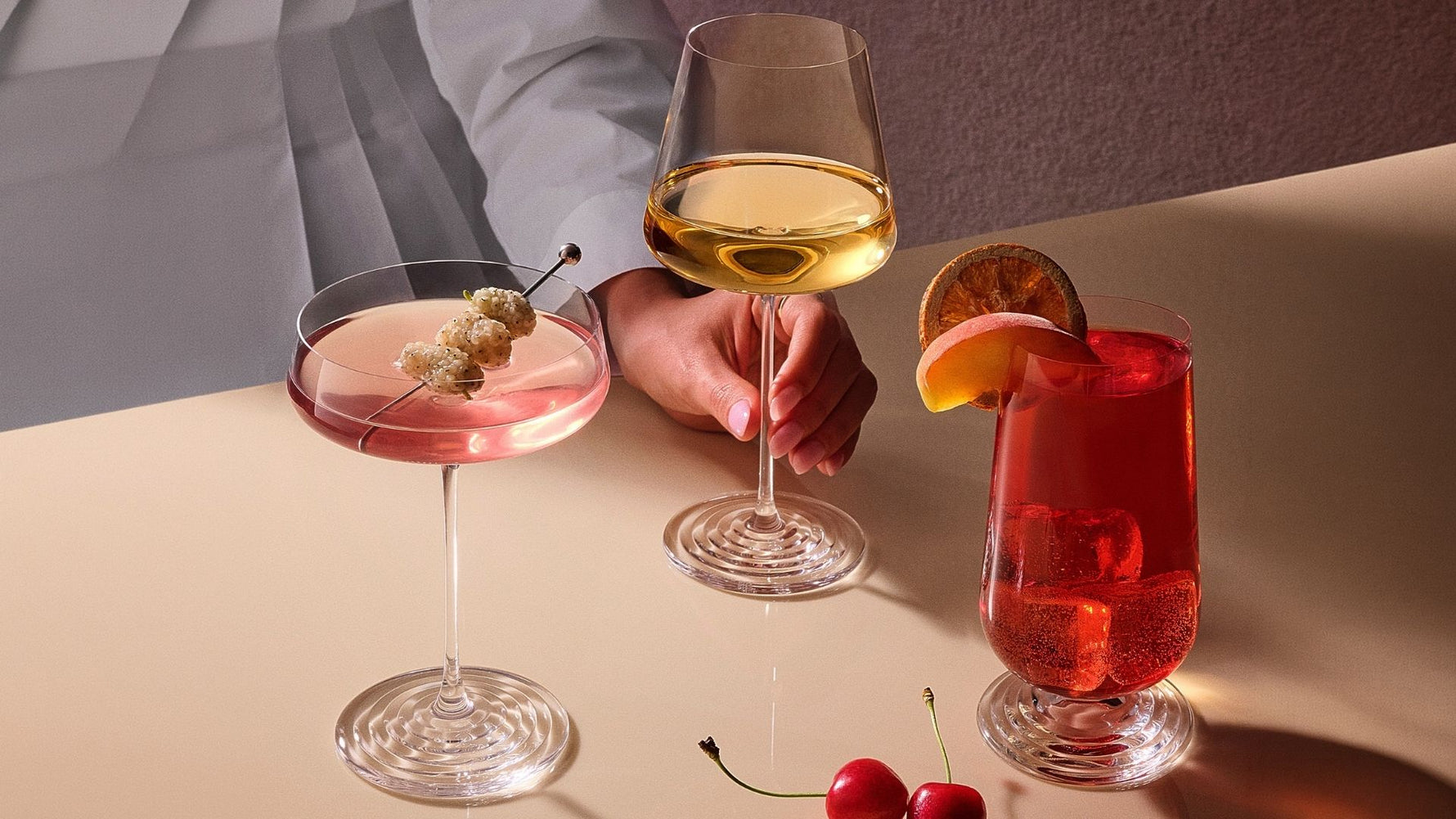 NUDE Splash by Tom Dixon glasses — Champagne Coupe with a pink cocktail, White Wine Glass, and Highball with a red spritz — on a warm-toned table.