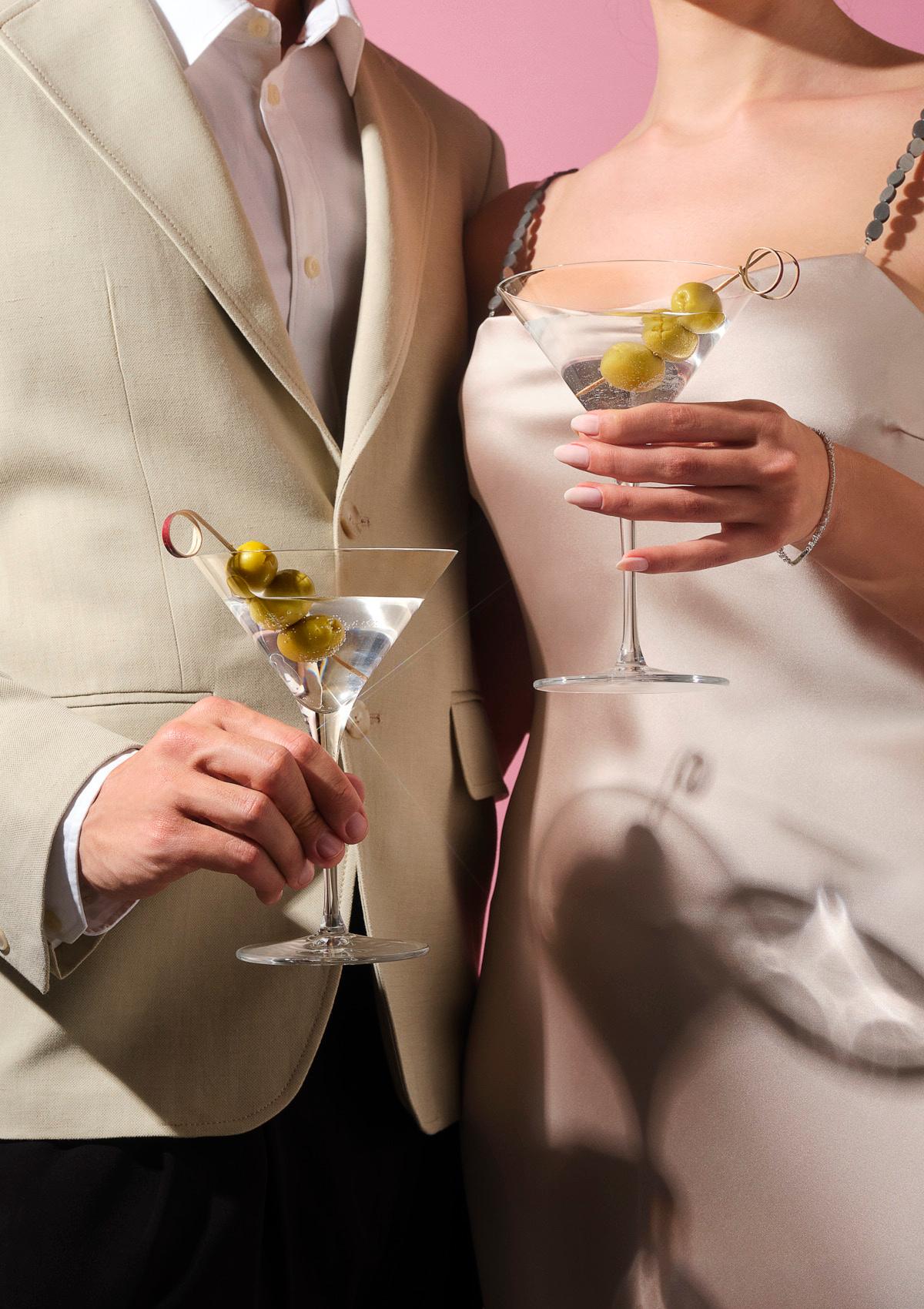 A couple in a linen suit and silk slip dress holding two NUDE Vintage martini glasses with olives.