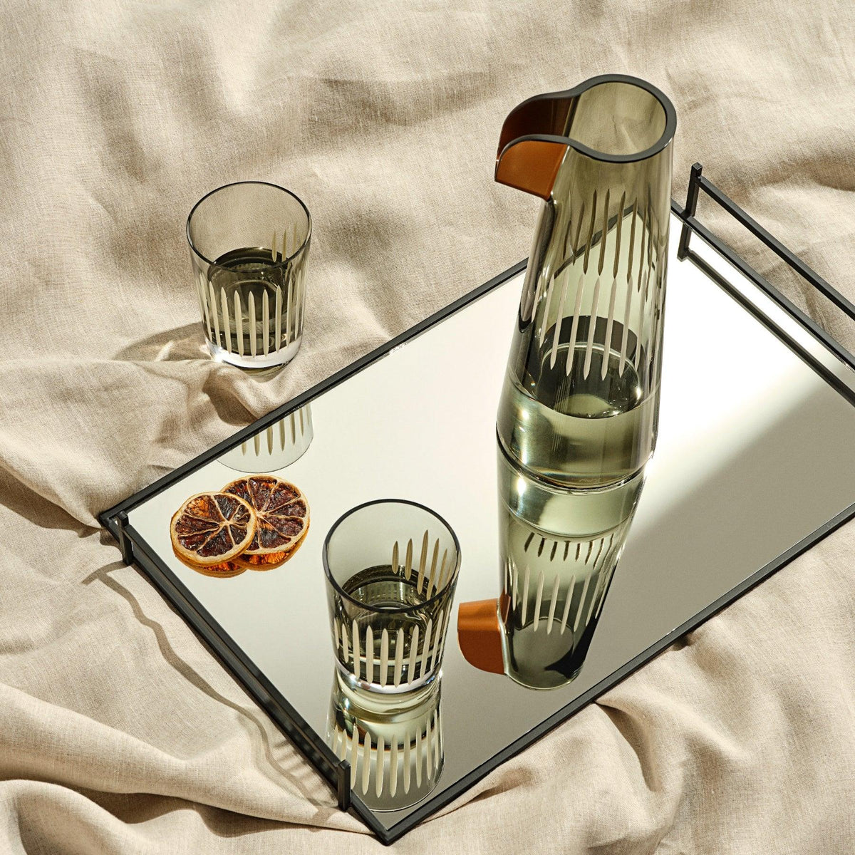 A clear crystal carafe and two tumblers with a geometric pattern, displayed on a reflective surface, accompanied by dried orange slices and a beige textile backdrop.