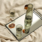 A clear crystal carafe and two tumblers with a geometric pattern, displayed on a reflective surface, accompanied by dried orange slices and a beige textile backdrop.